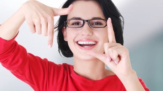 A young woman with black hair and glasses and perfect white teeth frames her face with her fingers.