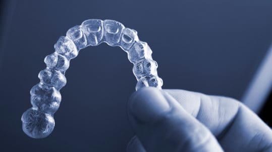 Light illuminates an invisalign retainer that's being held between thumb and forefinger over a dark blue background.