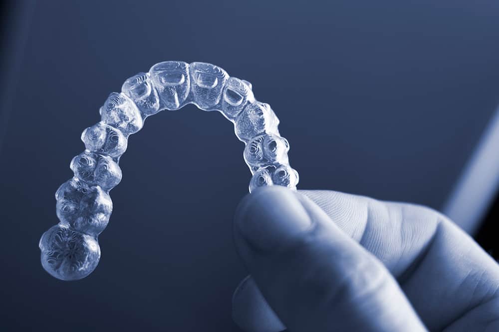 Light illuminates an invisalign retainer that's being held between thumb and forefinger over a dark blue background.