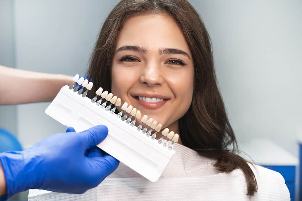A blue glove holds a case of sample dental veneers in front of a smiling girl with dark brown hair.