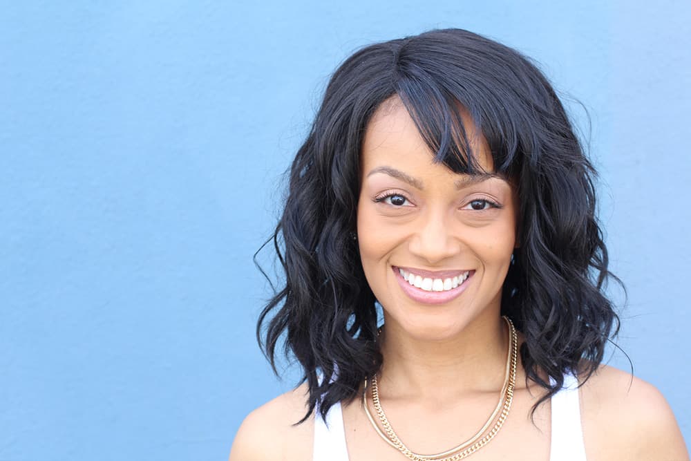 An attractive black woman with straight hair and white teeth smiles in front of a light blue background.