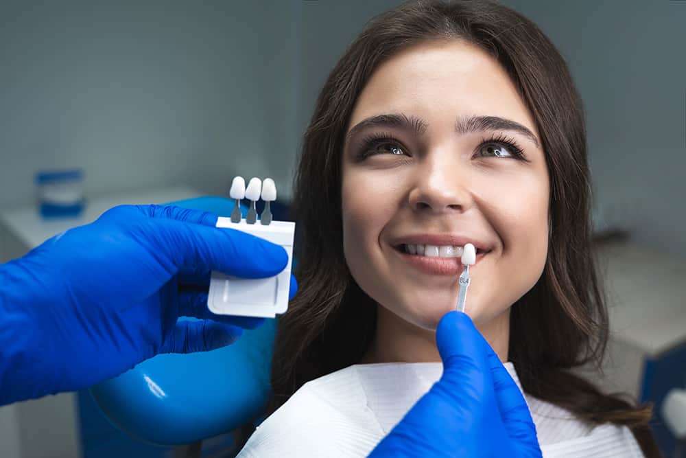 A young woman smiles at the dentist as hands wearing blue gloves hold dental veneers up to her teeth. 