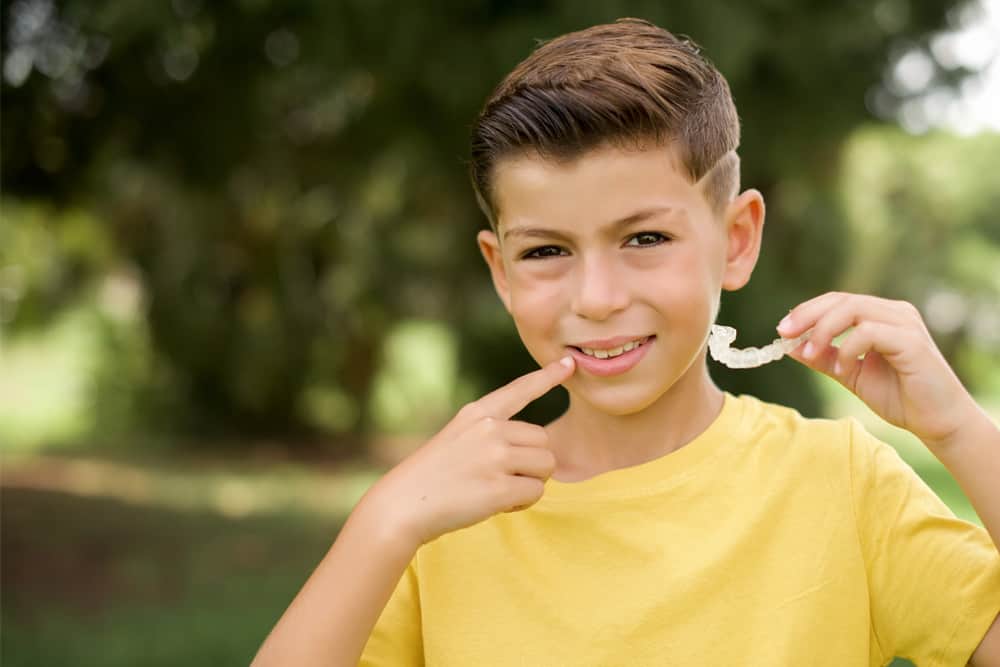 A young boy in a yellow shirt points to his smile while holding his invisalign retainer up by his smile. 