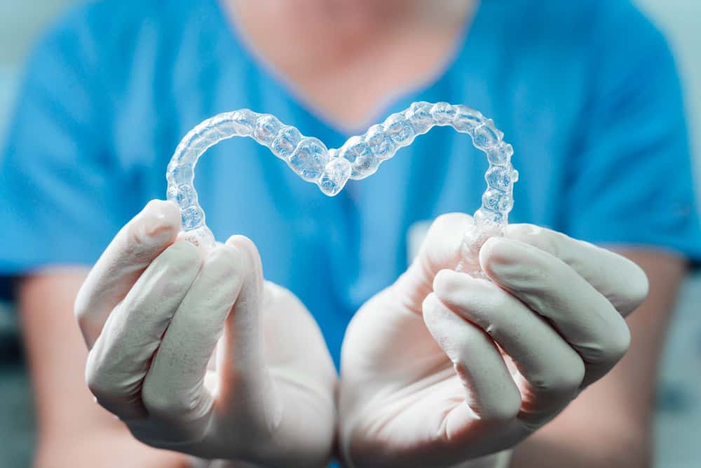Two invisalign retainers held together so that they form a heart with the gloved hands of the dentist. 