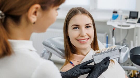 A woman smiles at the camera as a female dentist in gloves holds up veneers for her to see.