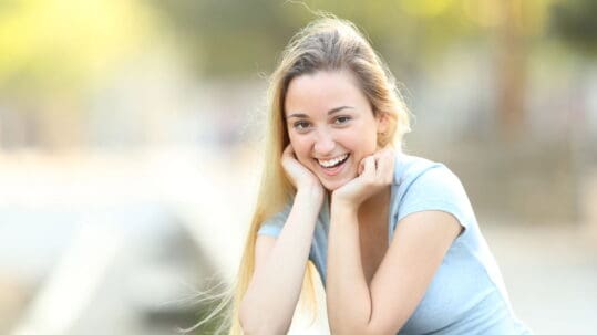 A young woman in a light blue shirt smiles at the camera in front of a blurred natural background.