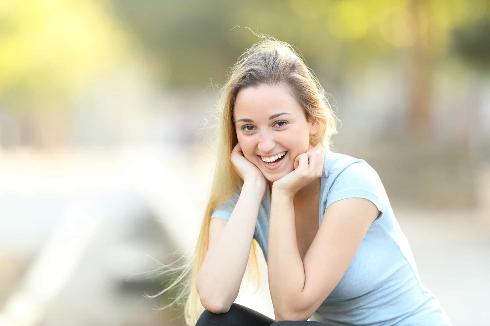 A young woman in a light blue shirt smiles at the camera in front of a blurred natural background.