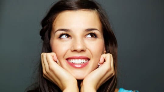 A young woman with perfect teeth smiles off screen in front of a dark gray background.