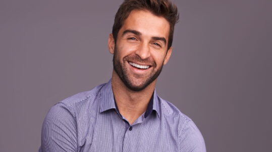 A man smiles to show off his porcelain veneers in front of a dark purple background.