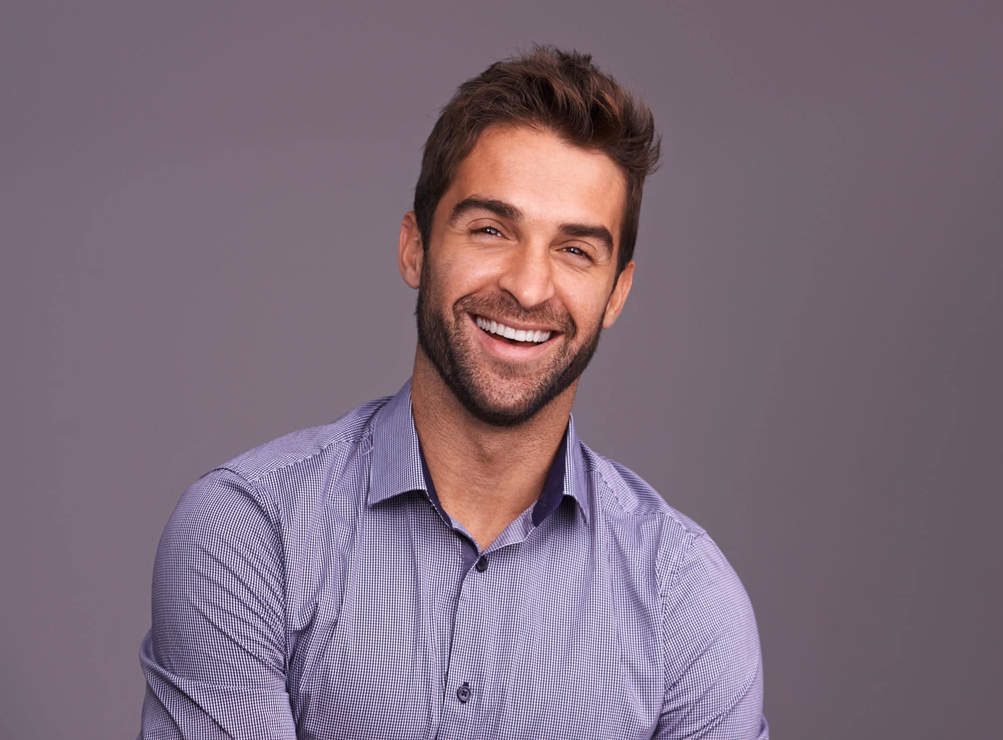 A man smiles to show off his porcelain veneers in front of a dark purple background.