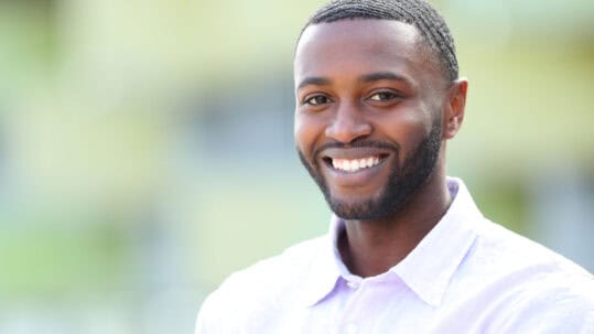 An attractive black man with perfect teeth smiles at the camera in front of a blurred background.