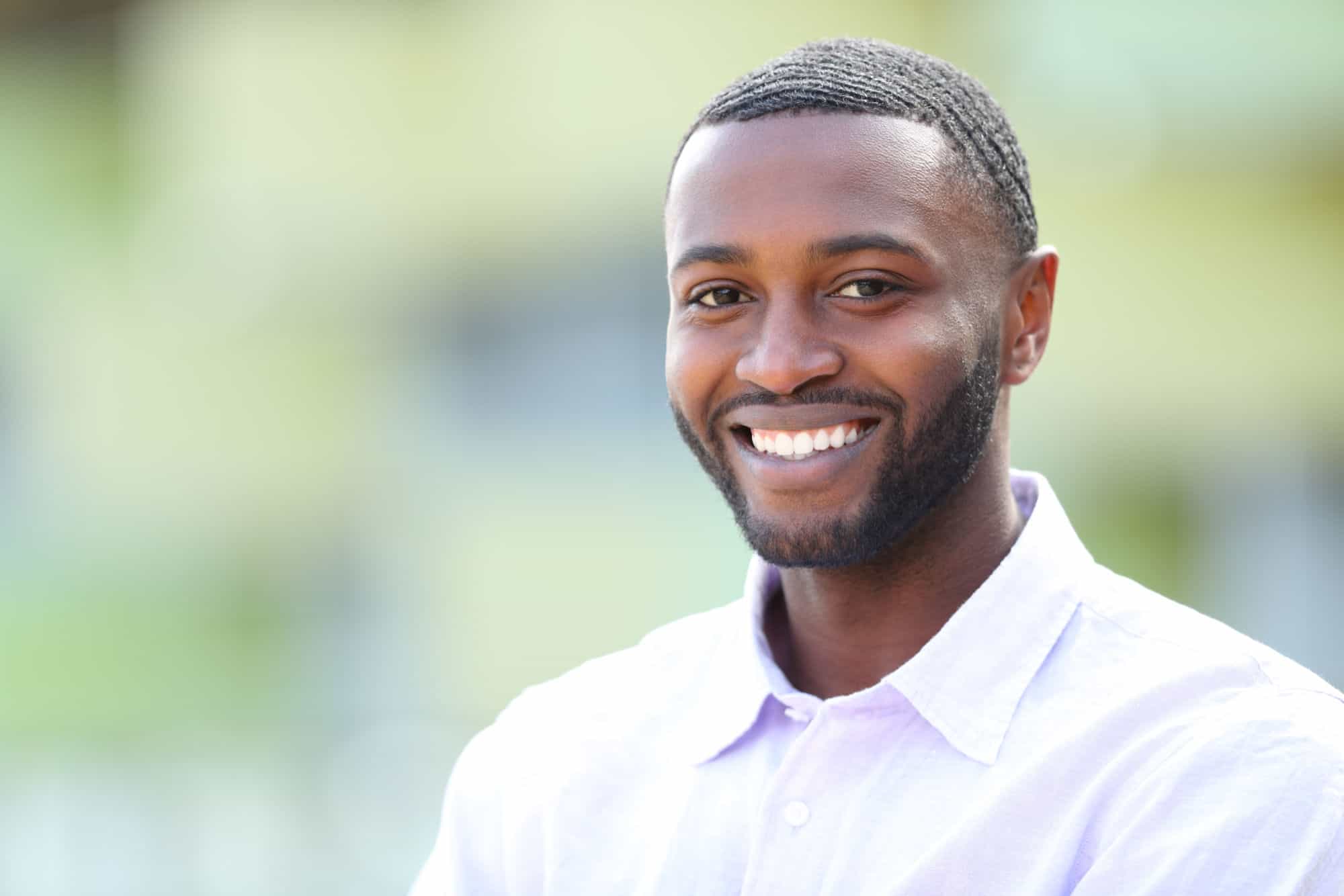 An attractive black man with perfect teeth smiles at the camera in front of a blurred background.