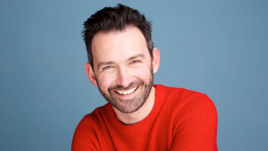 A handsome man with perfect teeth and a red sweater smiles at the viewer in front of a blue background.