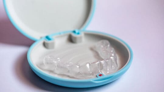 An Invisalign retainer in a blue case in front of a light pink background.