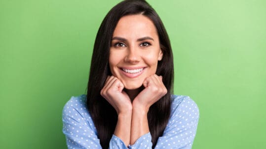 A beautiful woman with dark brown hair shows off her perfect smile after cosmetic dentistry in front of a green background.