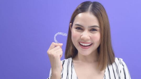 A young woman holds up her invisalign retainer while smiling in front of a purple background.