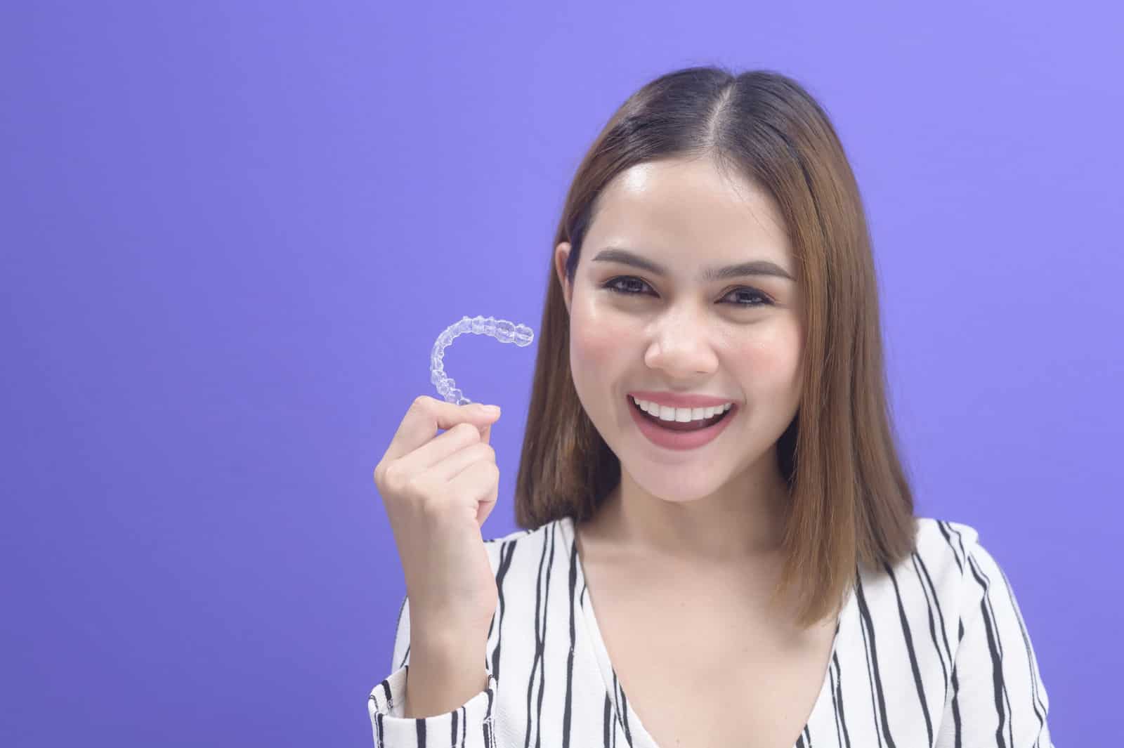 A young woman holds up her invisalign retainer while smiling in front of a purple background.